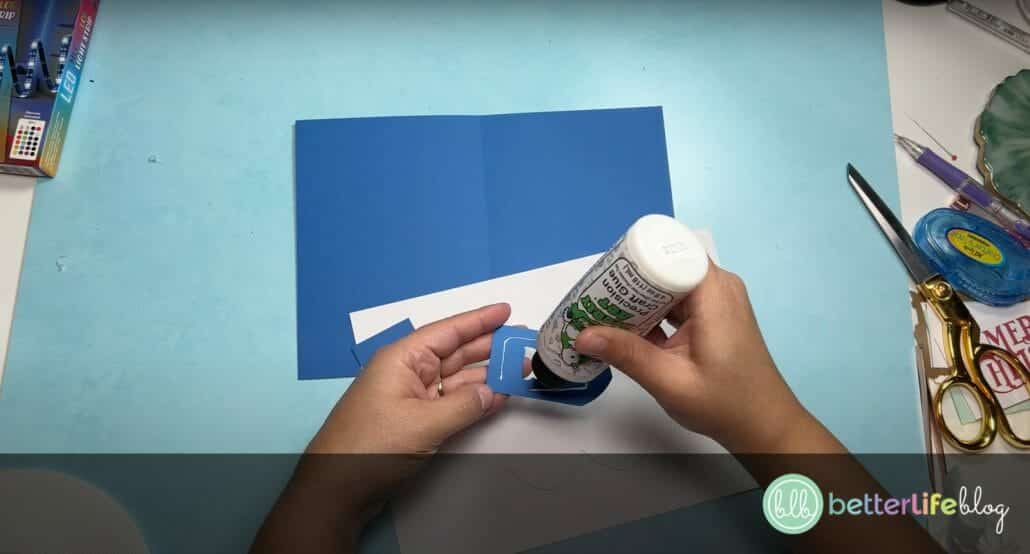 Adding glue to a small blue cardstock piece, with blue and white cardstock lie in the background